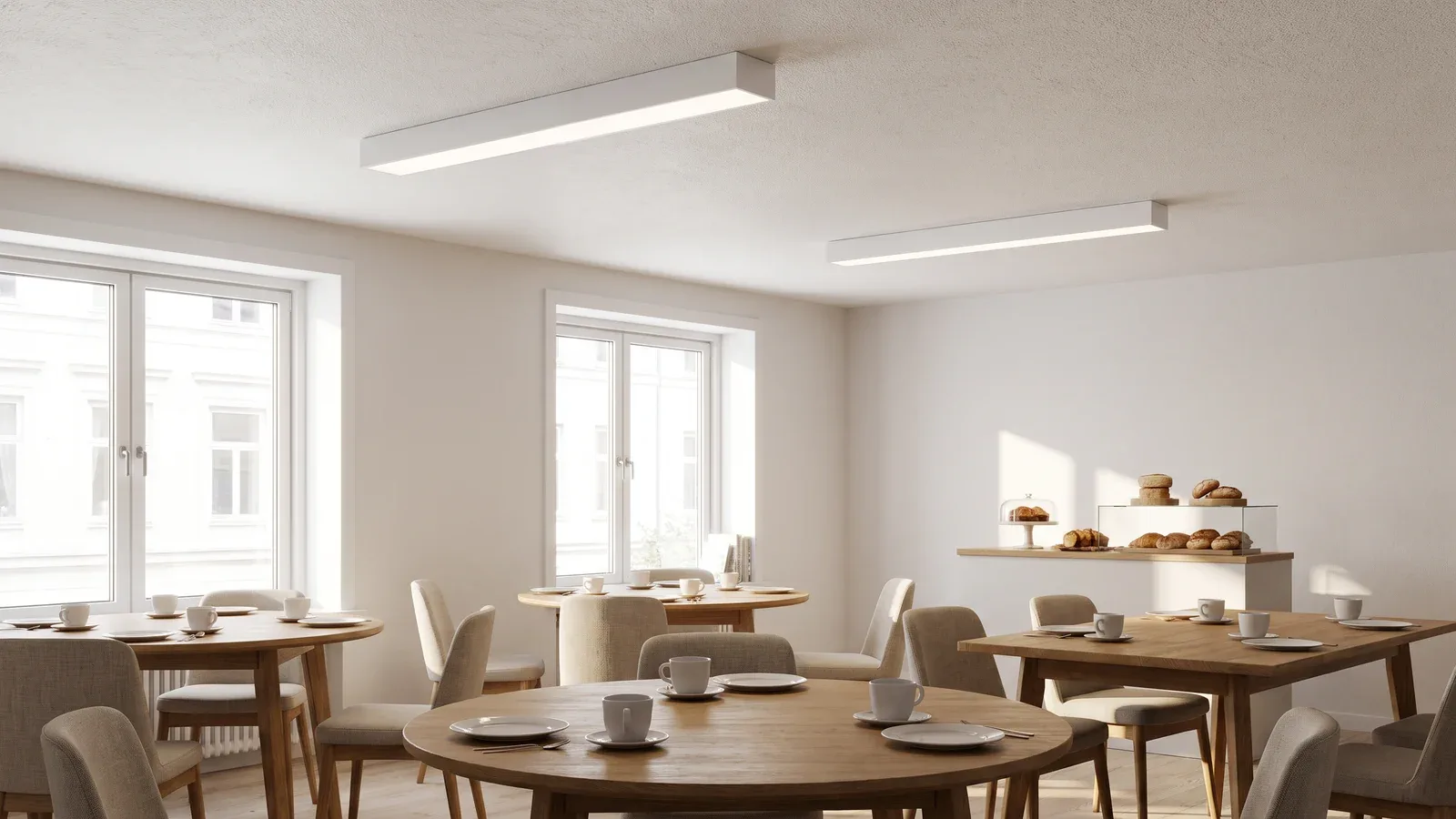 Salon de petit-déjeuner d’hôtel avec luminaires linéaires blancs en applique au plafond au-dessus des tables et lumière naturelle.